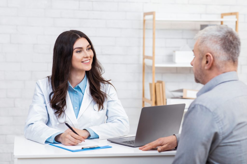 Beautiful doctor in white medical coat listening to old patient and making notes in tablet